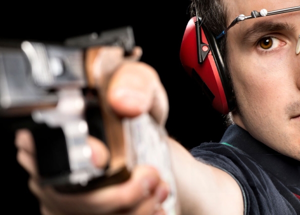 Um rapaz com tapa olho, abafador de ruído no ouvido e arma na mão, em posição de atirar