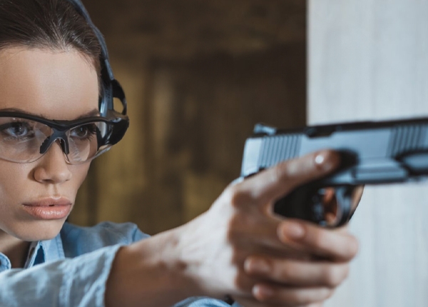 Uma mulher com óculos de proteção, abafador de ruído e arma nas mãos, em posição de atirar